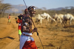 Ein Samburu-Krieger in traditioneller Kleidung trägt einen Speer, im Hintergrund sind weitere Menschen sowie Kamele zu erkennen.