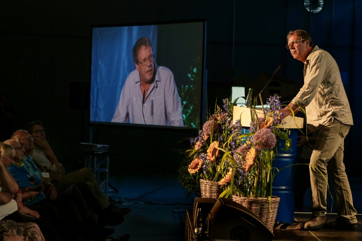 Georg Schramm steht auf der Bühne, links im Hintergrund die Video-Übertragung.
