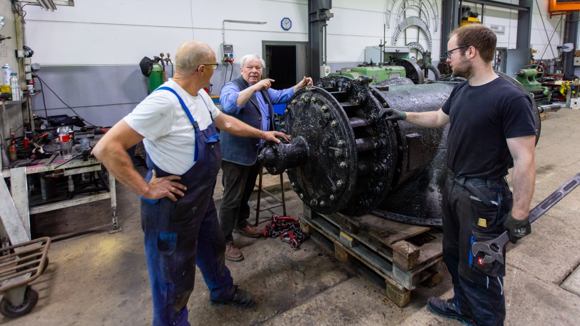 Drei unterschiedlich alte Männer in Arbeitskleidung werkeln an einer Turbine und unterhalten sich dabei.