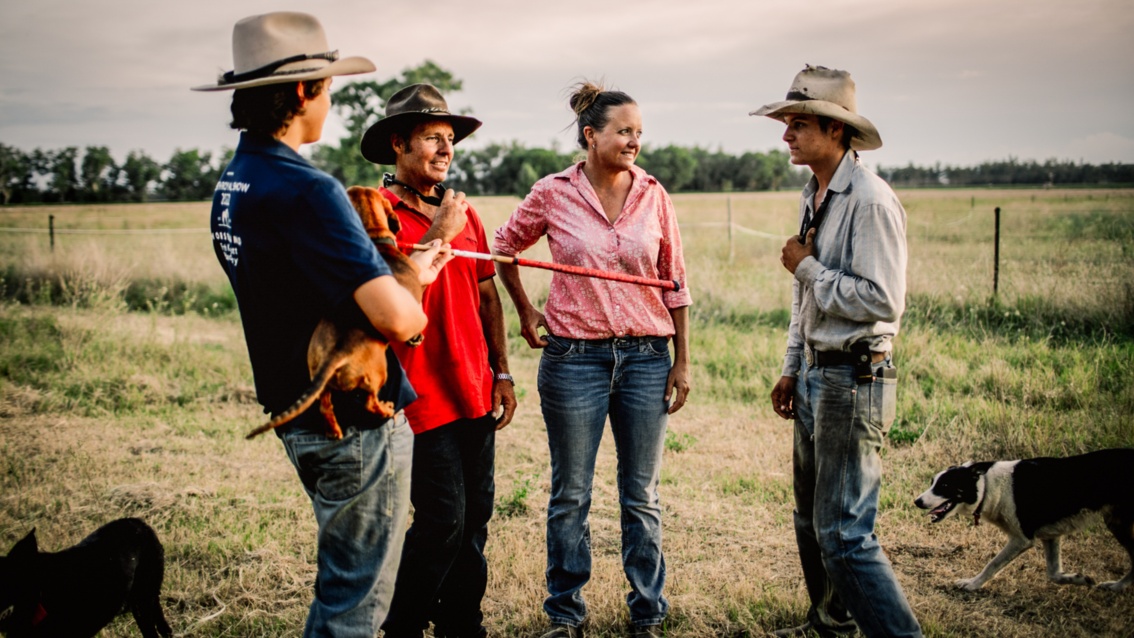 Sally, ihr Mann und die beiden Söhne, alle Männer mit Cowboyhüten stehen im Gespräche auf einer Weide, um sie herum hre beiden Hunde.