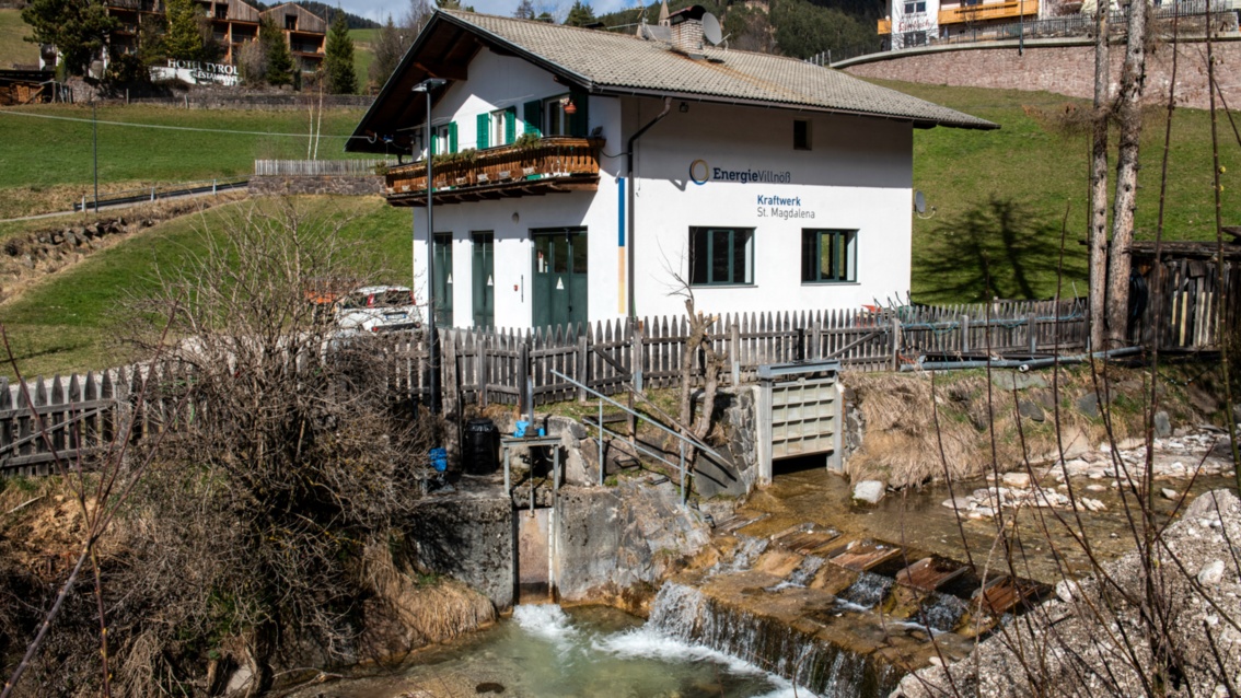 Ein kleines Wasserkraftwerk steht neben einem Gebirgsbach, hinter dem unscheinbaren Gebäude erstreckt sich eine Wiese.