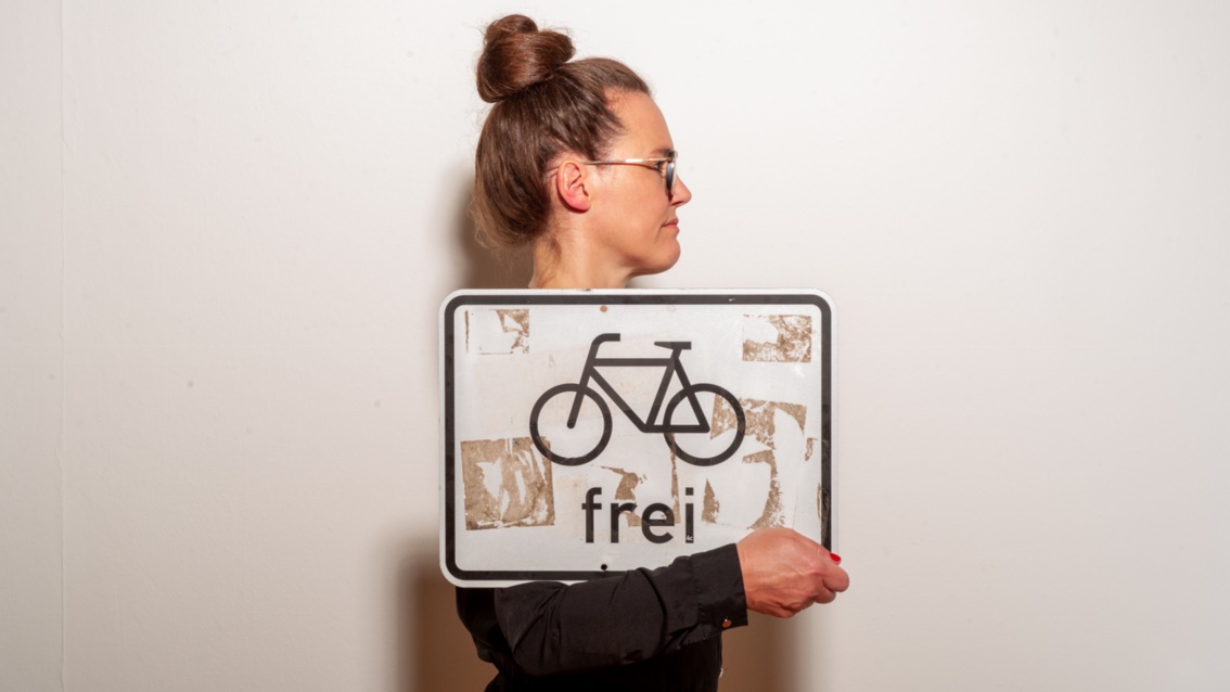 Nun steht die Frau mit dem Profil zur Kamera – seitlich klemmt ein „Radfahrer frei“ Schild in ihrem Arm. 